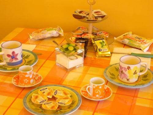 A cozy indoor dining setup features a vibrant yellow-orange checkered tablecloth adorned with colorful teacups, pastries, and a tiered tray of treats, creating a cheerful and inviting atmosphere perfect for a relaxing tea party or breakfast gathering.