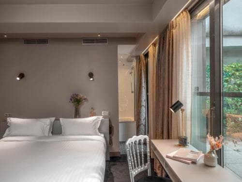 A cozy hotel room features a neatly made bed, a desk with a lamp, and a glass door leading to a serene outdoor garden, ideal for travelers seeking relaxation and comfort.
