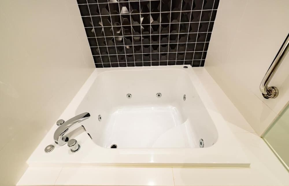A modern bathroom features a sleek white corner jacuzzi tub with chrome fixtures against a striking black geometric tile accent wall, ideal for travelers seeking relaxation in a stylish hotel suite.