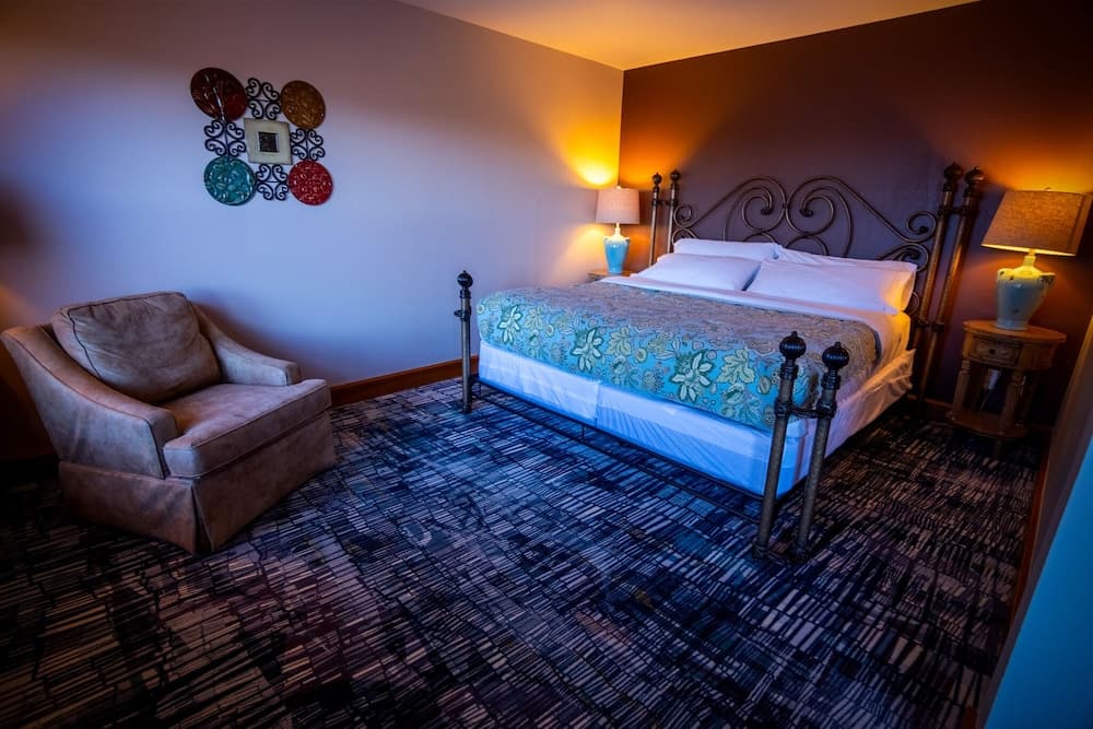 A cozy hotel room features a king-sized bed with ornate metal framing, vibrant bedding, ambient lighting from bedside lamps, a plush armchair, and colorful wall art, ideal for travelers seeking comfort and relaxation.