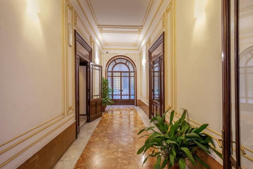 A serene hotel corridor features elegant gold-trimmed walls, polished marble floors, lush green plants, and an arched window at the end, ideal for travelers seeking refined accommodations.
