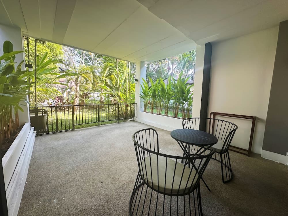 A serene balcony with a cozy black table and chairs overlooks lush greenery through large windows, ideal for travelers seeking a tranquil retreat in a tropical setting.