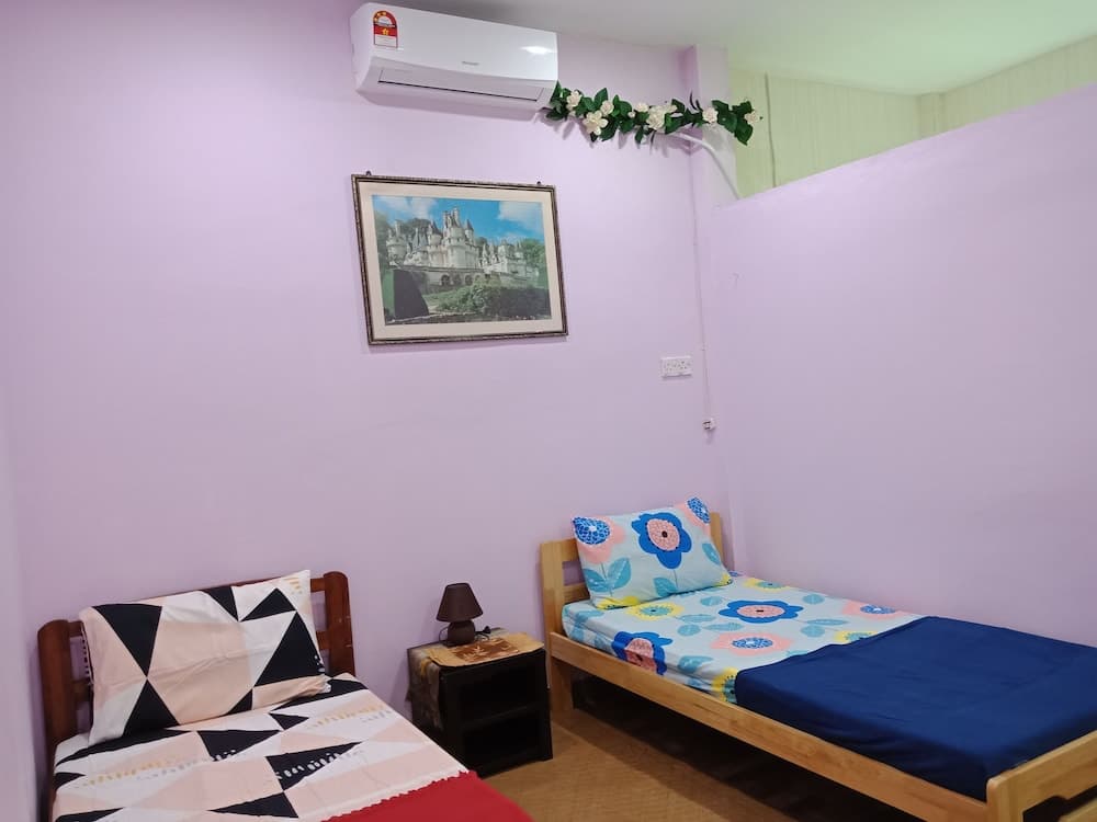 A cozy guesthouse room with lavender walls, two single beds adorned with colorful bedding, a framed castle artwork, and an air conditioner, ideal for travelers seeking a comfortable and budget-friendly stay.