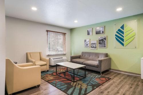 A cozy hotel lounge features modern furnishings, a vibrant rug, and a green accent wall adorned with artwork, creating a welcoming atmosphere ideal for travelers seeking relaxation.