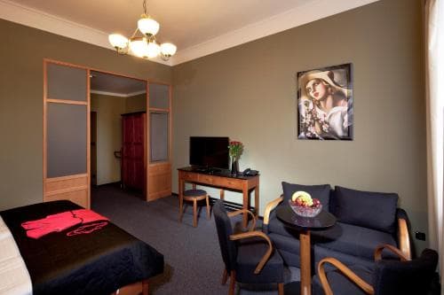 A cozy hotel room features a neatly made bed, a small dining area with a fruit bowl, vintage-style furniture, and warm lighting, creating an inviting atmosphere ideal for travelers seeking comfort and relaxation.