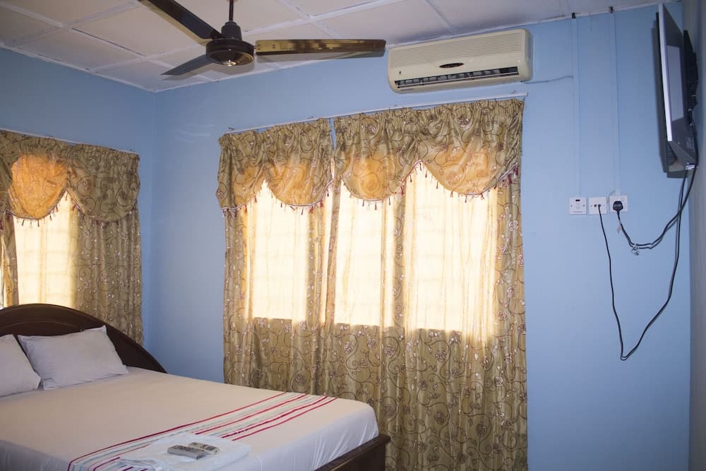 A cozy hotel room with blue walls, floral curtains, a neatly made bed, air conditioning, and a mounted TV offers a comfortable stay for travelers seeking relaxation.