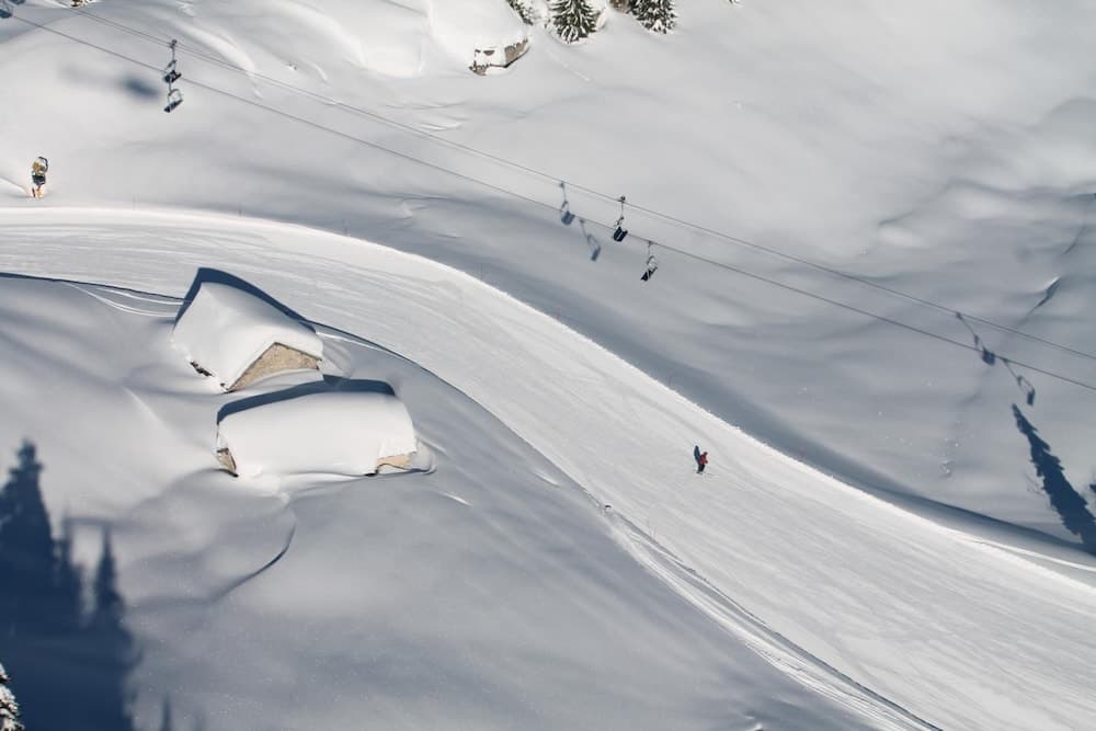 A serene winter landscape features a snow-covered ski slope with skiers gliding down, a cable car system overhead, and quaint snow-laden cabins nestled in the mountains, ideal for adventure seekers planning a ski vacation.