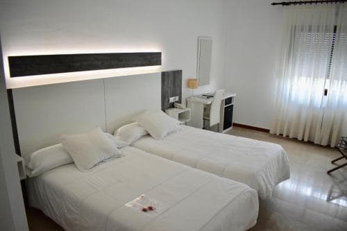 A cozy hotel room features two neatly made beds under soft lighting, a desk area by the window with sheer curtains, and a minimalist design, ideal for travelers seeking comfort and simplicity.
