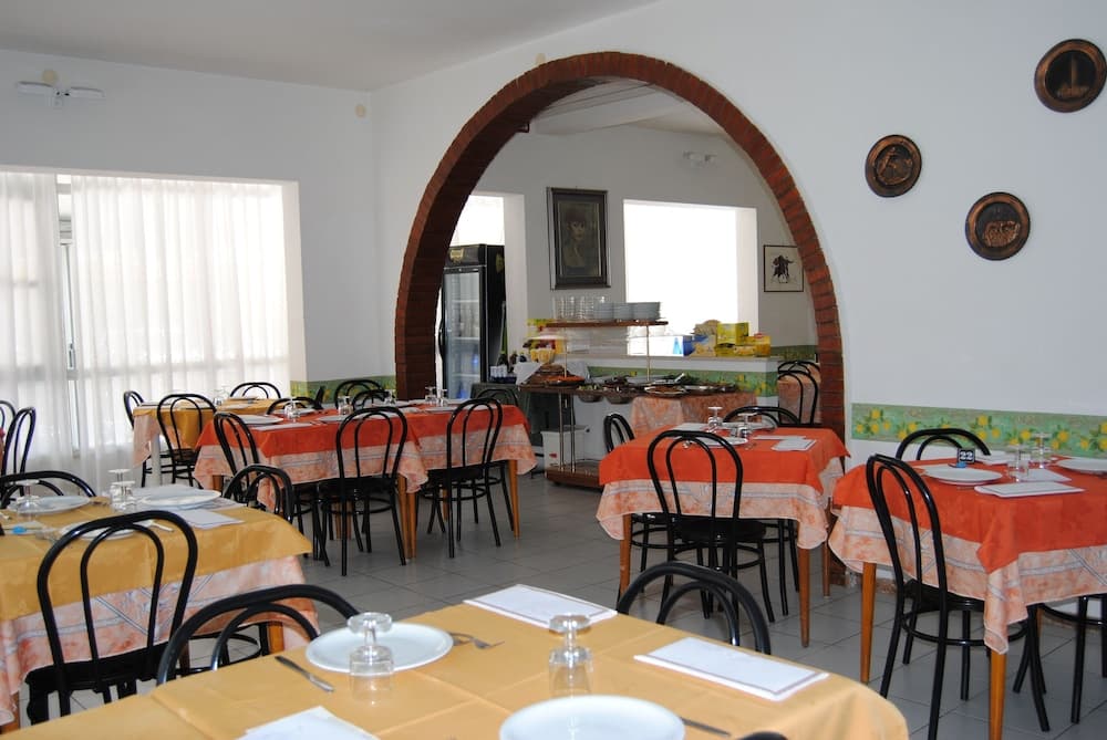 A cozy restaurant interior features tables draped in orange tablecloths under a tiled floor, framed by a brick archway leading to a bright kitchen area, ideal for travelers seeking a warm dining experience.