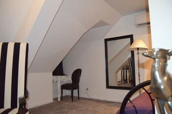 A cozy attic room with slanted ceilings features a stylish black-and-white striped chair, a vanity table with a lamp, and a large mirror, creating an intimate and elegant space ideal for personal retreats or boutique hotel stays.