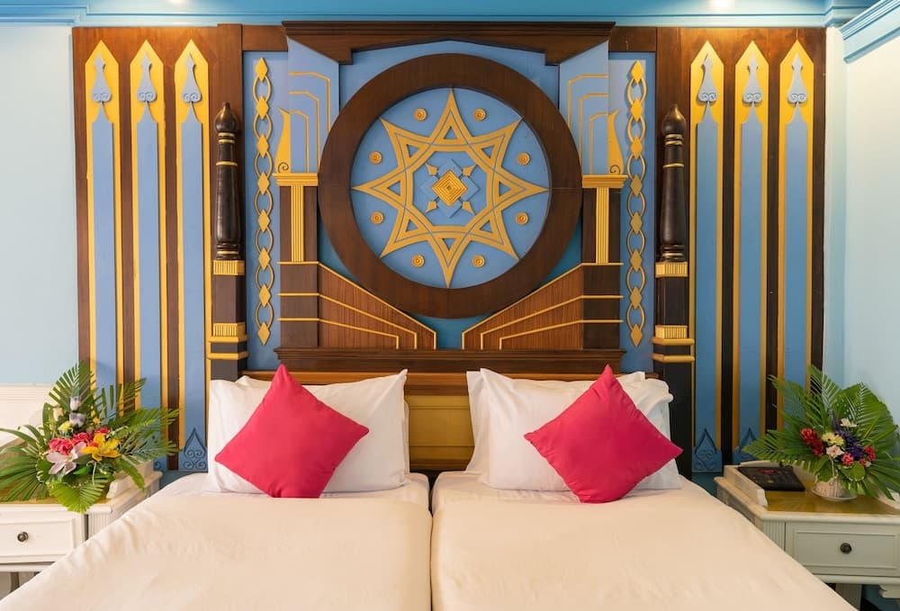 A vibrant hotel room features an ornate wooden headboard with intricate gold and blue designs, complemented by red pillows and lush floral arrangements on white bedside tables, ideal for travelers seeking a luxurious and culturally rich accommodation experience.