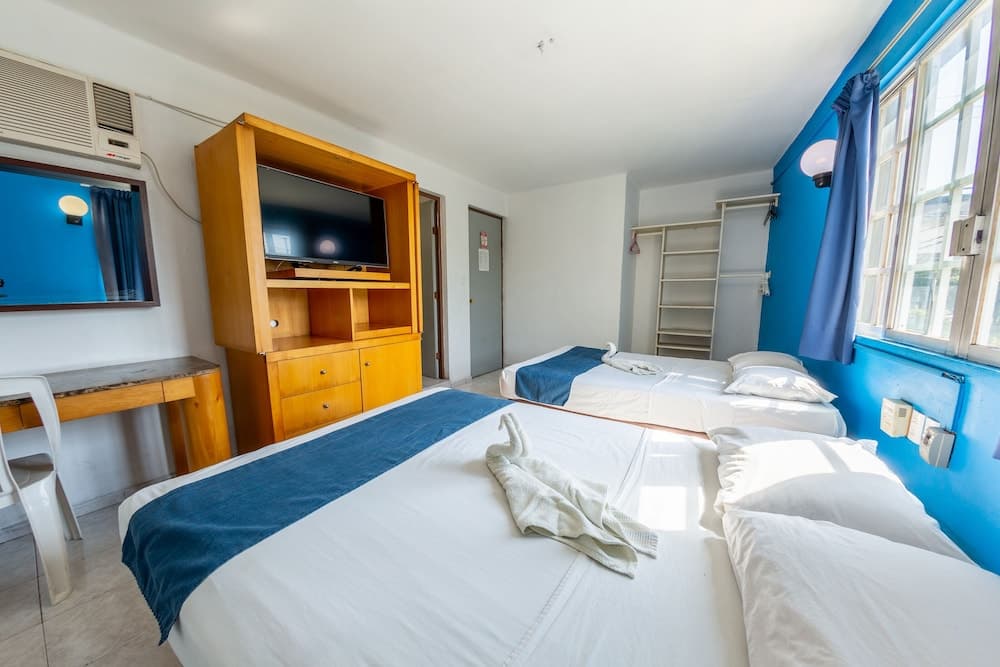 A cozy hotel room with two neatly made beds, blue accents, a wooden TV cabinet, and ample natural light through large windows, ideal for travelers seeking comfort and simplicity.