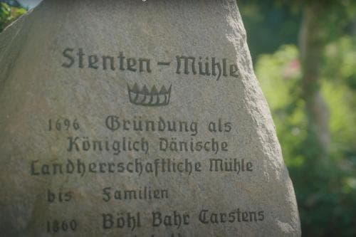 A serene stone monument inscribed with "Stenten-Mühle" stands amidst lush greenery, commemorating its historical significance as a Danish royal mill since 1696, ideal for cultural heritage tourism.