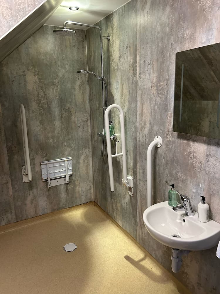 A modern, accessible bathroom with textured walls, a walk-in shower featuring grab bars and a fold-down seat, a wall-mounted sink, and a mirror, ideal for travelers seeking comfort and inclusivity in accommodations.