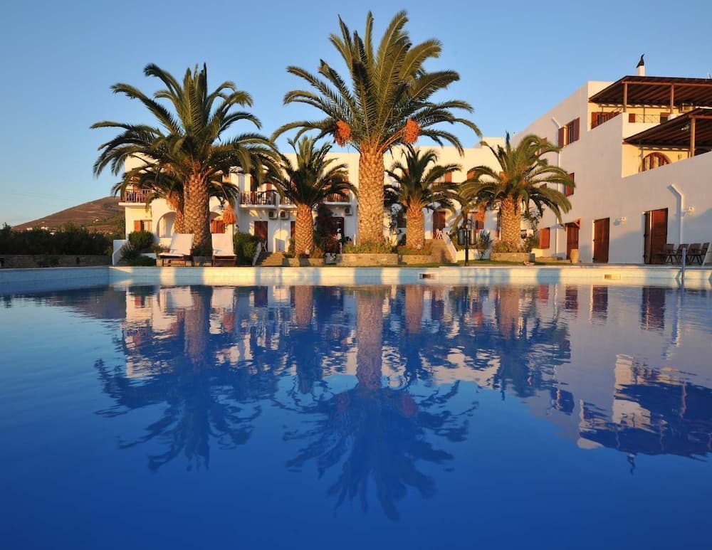 A serene resort scene features a tranquil pool reflecting palm trees and white buildings under a clear sky, ideal for vacation relaxation.