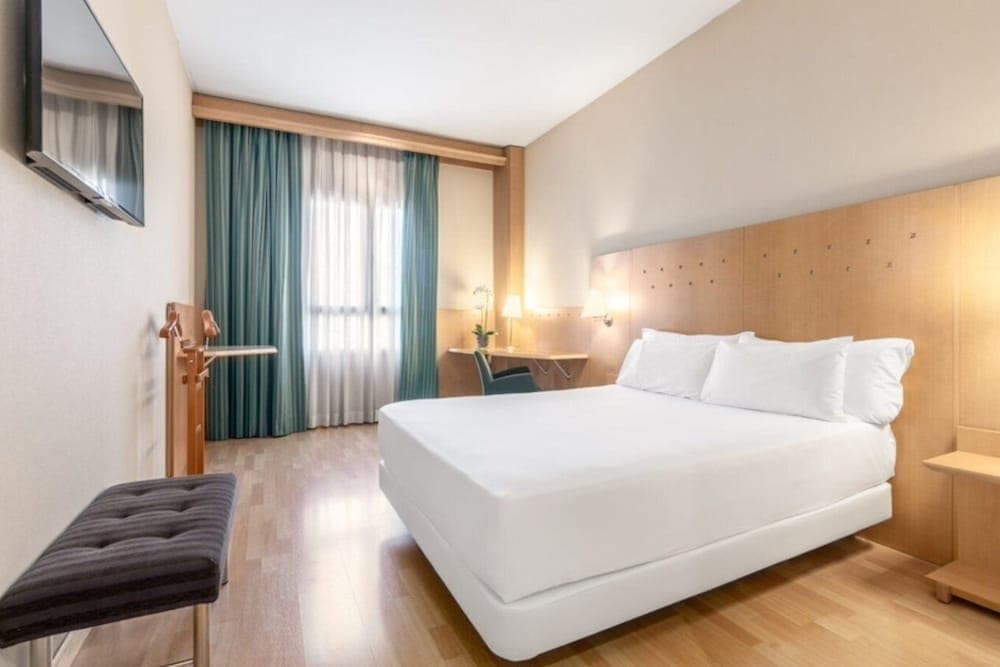 A cozy hotel room features a neatly made bed with white linens, wooden furnishings, a desk by the window with sheer curtains, and a wall-mounted TV, ideal for travelers seeking comfort and functionality.