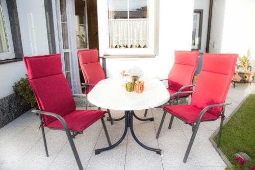 A cozy outdoor patio features vibrant red chairs around a white table set with colorful fruits, inviting travelers to relax in a serene residential setting.