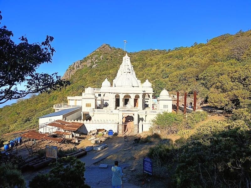 A serene view of the Jal Mandir temple nestled against lush green hills under a clear blue sky, featuring intricate white architecture and surrounded by natural beauty, ideal for spiritual retreats and cultural exploration in Jharkhand, India.