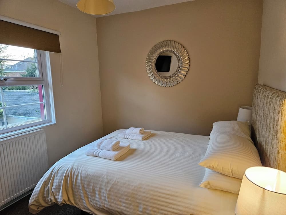 A cozy bedroom with neatly folded towels on the bed, a sunburst mirror on the wall, soft lighting from bedside lamps, and a window view of rooftops, ideal for travelers seeking a serene and comfortable stay.