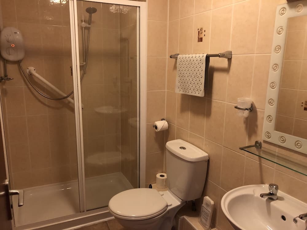A cozy hotel bathroom features beige tiled walls, a glass-enclosed shower, a toilet with dual paper rolls, a sink with a decorative mirror, and a towel rack with a textured towel, ideal for travelers seeking comfort and functionality.