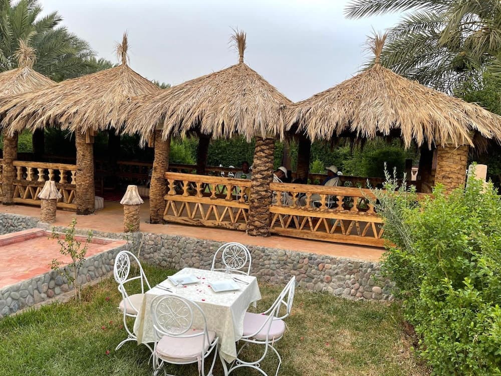 A serene outdoor dining area features rustic thatched-roof pavilions surrounded by lush greenery, creating an inviting atmosphere perfect for travelers seeking a tranquil retreat.