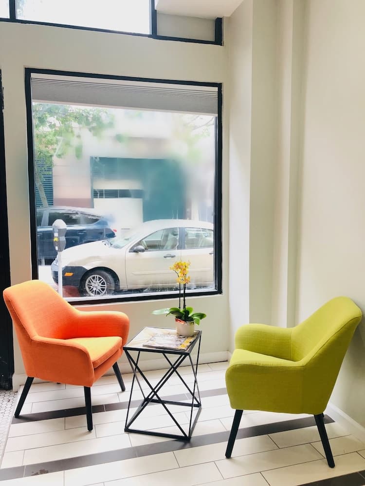 A cozy indoor seating area features vibrant orange and green chairs flanking a small table with a potted plant, set against a large window offering a view of parked cars outside, ideal for travelers seeking a relaxing spot in a modern urban setting.