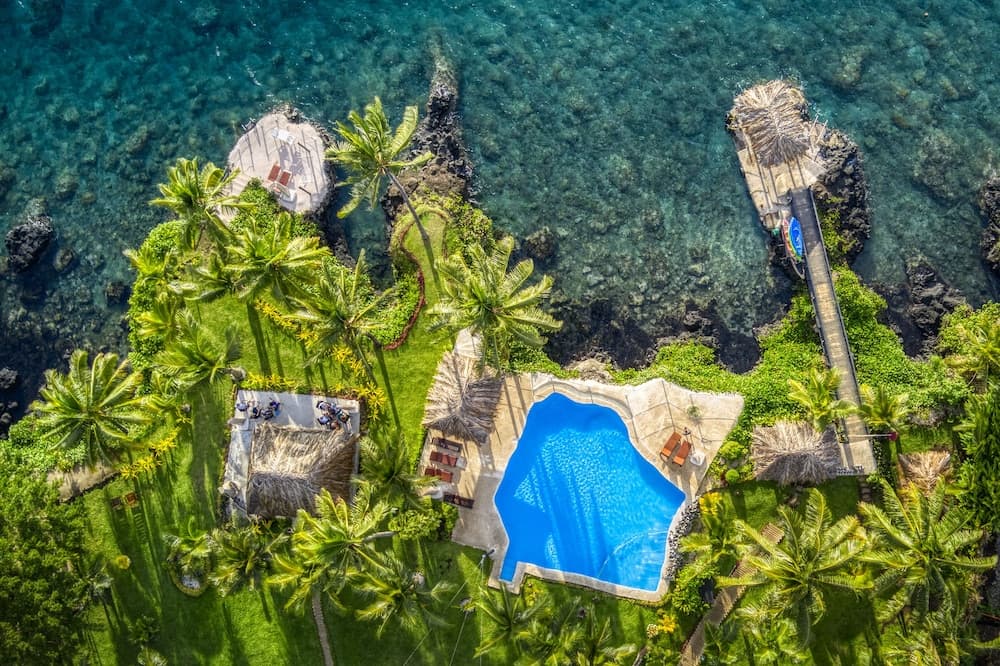 An aerial view of a luxurious tropical resort with lush greenery, palm trees, a vibrant blue pool, and a wooden pier extending into crystal-clear turquoise waters, ideal for travelers seeking a serene and exotic getaway.