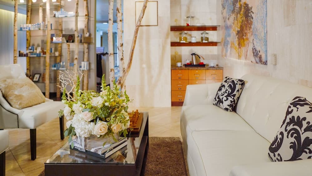 A cozy spa lounge features plush white sofas adorned with patterned pillows, a vibrant floral centerpiece on a glass coffee table, and birch tree decor, creating a serene atmosphere ideal for relaxation and wellness retreats.