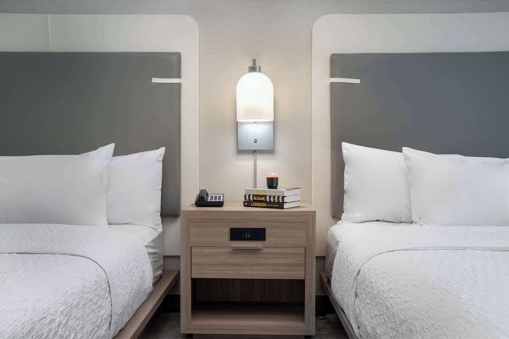 A cozy hotel room features two neatly made beds with white linens, a modern wooden nightstand holding books and a candle, and a soft-glowing wall-mounted lamp, ideal for travelers seeking comfort and relaxation.