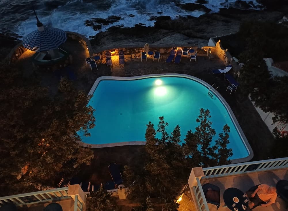 A serene night scene captures an illuminated pool surrounded by lounge chairs and a gazebo, overlooking the crashing waves of the ocean, ideal for a luxurious coastal retreat.