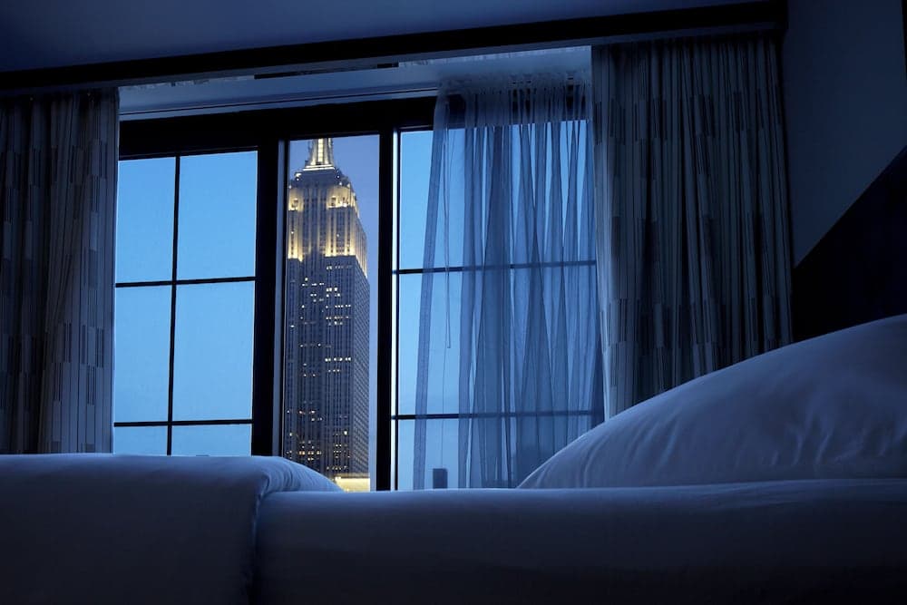 A cozy hotel room at dusk offers a breathtaking view of the illuminated Empire State Building through large windows, perfect for travelers seeking urban luxury and romance.
