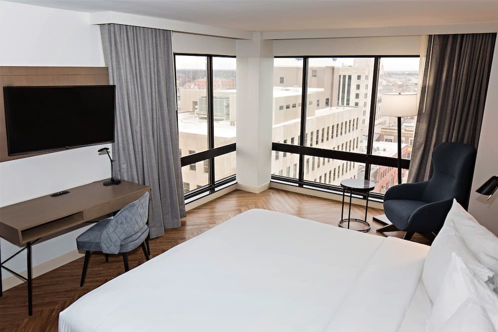 A cozy hotel room with a neatly made bed, modern desk setup, and large windows offering a panoramic city view, ideal for travelers seeking comfort and urban scenery.