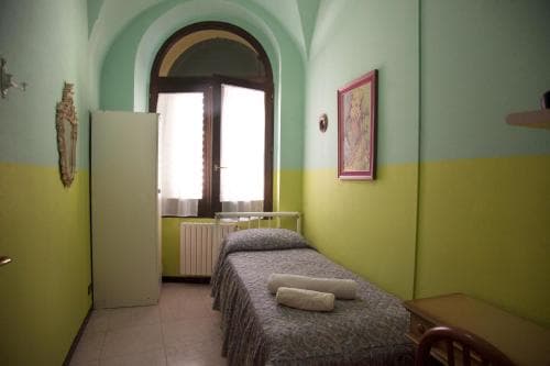 A cozy hostel room with vibrant green and yellow walls, featuring a neatly made bed, arched window, and simple furnishings, ideal for budget travelers seeking comfort in a charming setting.