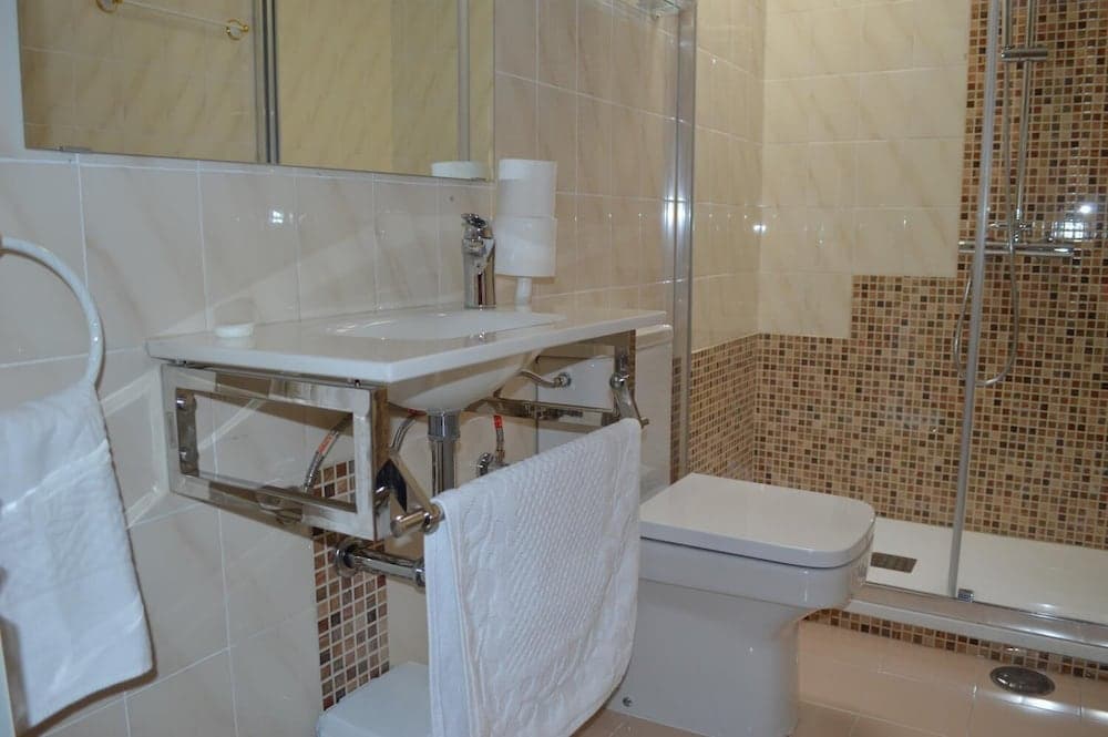 A cozy hotel bathroom features beige tiles, a modern sink with exposed plumbing, a glass-enclosed shower, and neatly hung white towels, ideal for travelers seeking comfort and cleanliness.