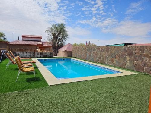 A serene backyard oasis features a rectangular pool surrounded by artificial turf, lounge chairs, and a stone wall, ideal for relaxing getaways under a partly cloudy sky.