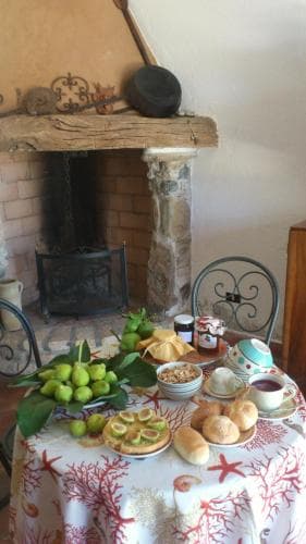 A cozy indoor dining setup features a rustic stone fireplace, a table adorned with fresh bread, fruits, and tea on a coral-patterned cloth, evoking a warm, inviting atmosphere perfect for a relaxing countryside retreat.