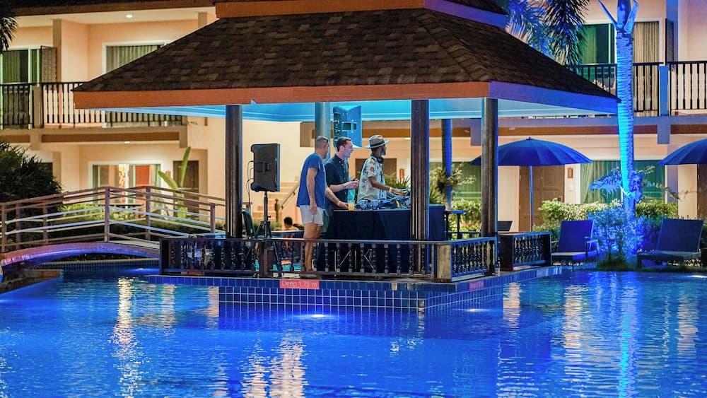A vibrant evening scene at a resort poolside area features a DJ booth under a gazebo, surrounded by lounge chairs and palm trees, creating an inviting atmosphere for travelers seeking relaxation and entertainment.