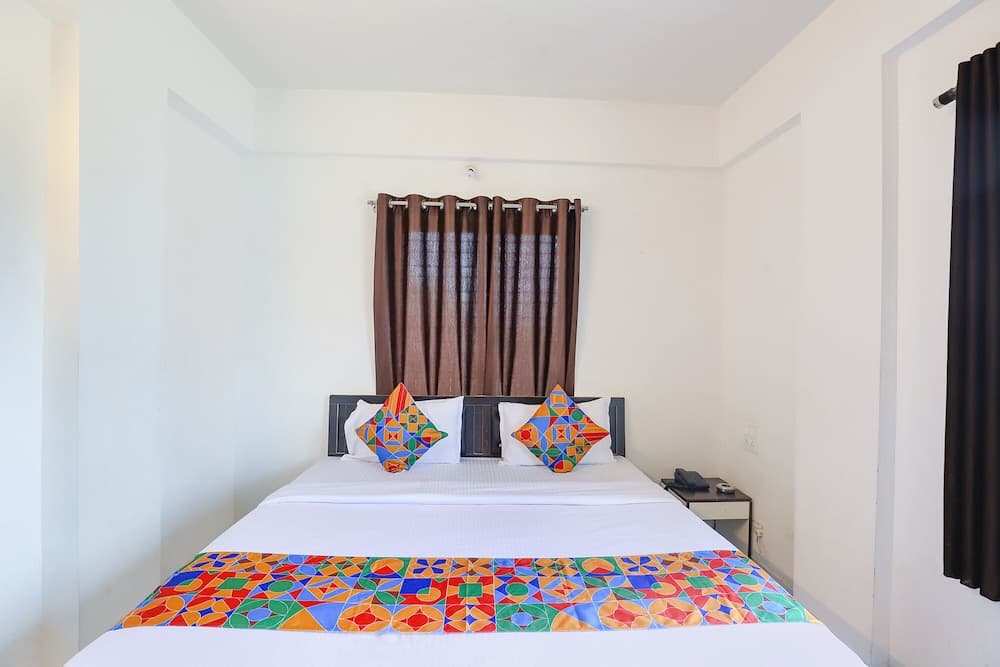 A cozy hotel room features a neatly made bed adorned with vibrant geometric-patterned pillows and a colorful throw, set against white walls and brown curtains, ideal for travelers seeking comfort and simplicity.