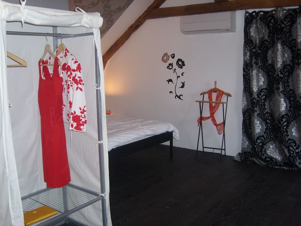 A cozy attic bedroom with exposed wooden beams, a simple bed, a clothing rack with vibrant red and floral garments, and elegant black-and-white curtains creates an intimate and stylish retreat ideal for travelers seeking a charming accommodation experience.