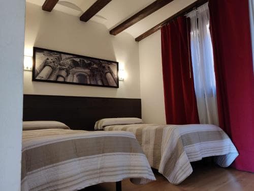 A cozy hotel room with twin beds, wooden beams, red curtains, and an architectural artwork creates a warm and inviting atmosphere ideal for travelers seeking comfort and charm.