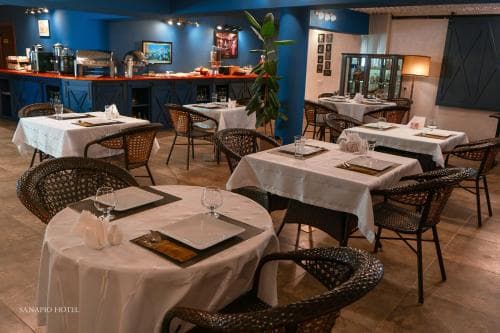 A cozy restaurant interior with blue walls, neatly arranged tables draped in white linens, wicker chairs, and warm lighting creates an inviting atmosphere ideal for travelers seeking a comfortable dining experience.