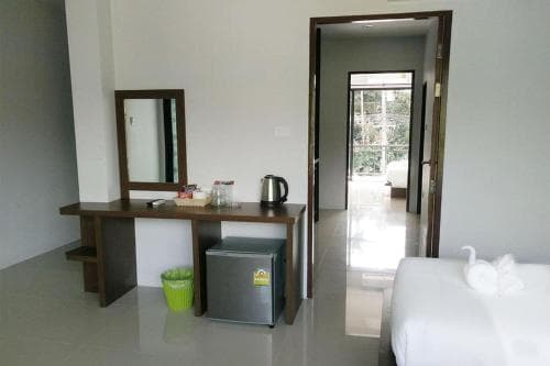 A cozy hotel room features a wooden vanity with a mirror, a mini-fridge, and a doorway leading to a balcony with lush greenery, ideal for travelers seeking comfort and relaxation.