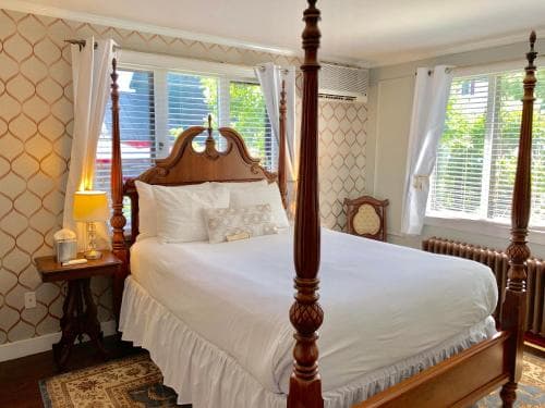 A cozy bedroom with a four-poster bed, patterned wallpaper, and soft natural light through large windows creates an inviting atmosphere ideal for a relaxing getaway at a boutique hotel.