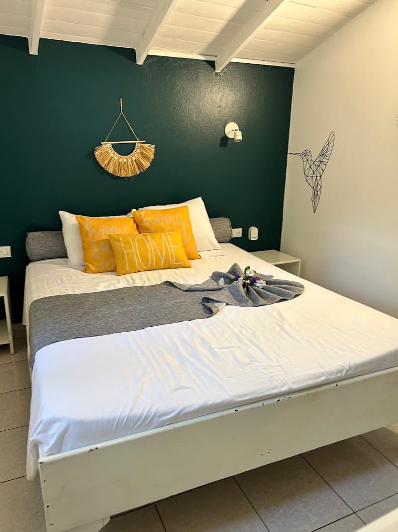 A cozy hotel room in Las Terrenas, Dominican Republic, features a neatly made bed adorned with yellow "HOME" pillows, a decorative wall hanging, and a hummingbird art piece, creating a welcoming atmosphere ideal for travelers seeking relaxation.