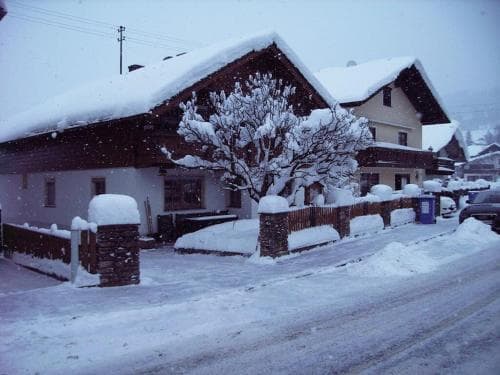 A serene winter scene captures a snow-covered house with a stone fence and tree, evoking a cozy alpine village atmosphere ideal for winter getaways.