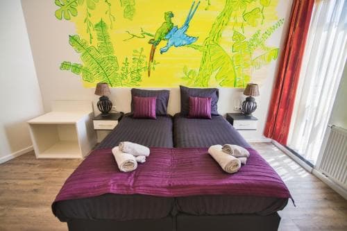 A cozy hotel room features twin beds adorned with purple bedding, vibrant tropical wall art, and natural light streaming through a window, ideal for travelers seeking comfort and a cheerful ambiance.