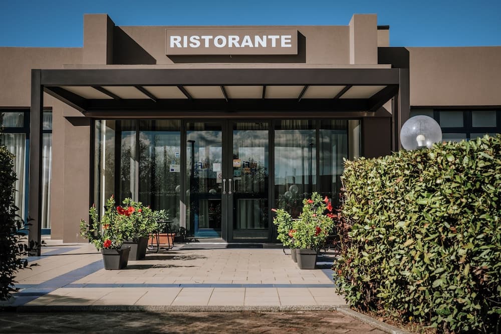 A welcoming ristorante entrance features modern glass doors framed by lush potted plants and a neatly trimmed hedge, ideal for travelers seeking an inviting dining experience under clear skies.