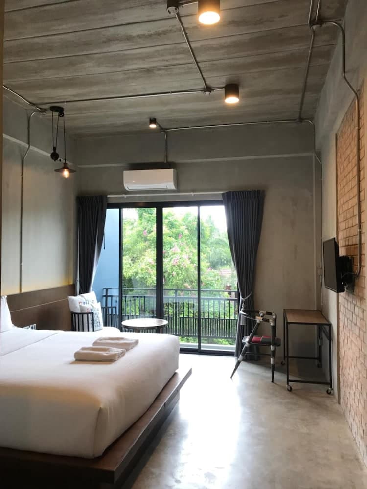 A cozy hotel room with an industrial chic design features exposed concrete ceilings, a brick wall, and large glass doors opening to lush greenery, ideal for travelers seeking a modern yet serene retreat.