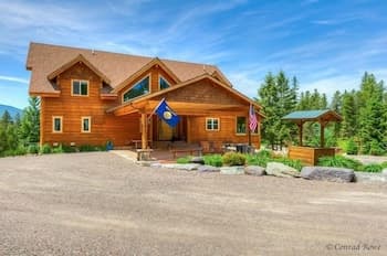 A serene mountain retreat features a cozy wooden cabin adorned with flags, surrounded by lush evergreens under a clear blue sky, ideal for a peaceful getaway.