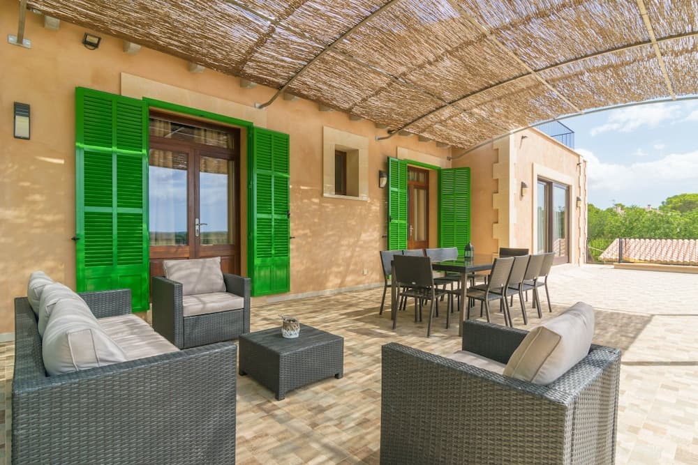 A serene outdoor terrace features comfortable wicker furniture under a thatched roof, set against a warm-toned building with vibrant green shutters, ideal for relaxing vacations in Mediterranean-style retreats.